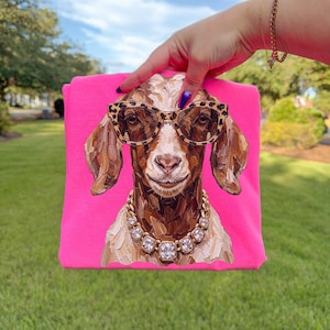 May include: A bright neon pink t-shirt featuring a central graphic of a brown goat wearing leopard print sunglasses and a chunky gold-toned necklace with clear faceted stones. The goat illustration has visible brushstroke textures.