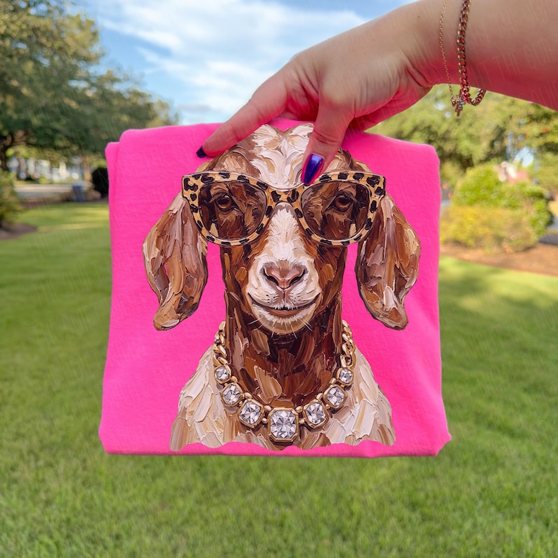 May include: A bright neon pink t-shirt featuring a central graphic of a brown goat wearing leopard print sunglasses and a chunky gold-toned necklace with clear faceted stones. The goat illustration has visible brushstroke textures.