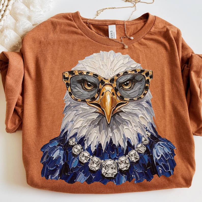 May include: A rust-colored t-shirt featuring a graphic of a bald eagle wearing leopard-print glasses and a crystal-style necklace. The eagle is rendered in a thick, textured paint style with white, gray, and blue feathers against the fabric.