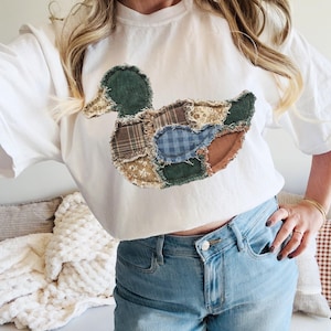 May include: A white short-sleeve t-shirt featuring a large duck-shaped applique on the chest. The duck is constructed from various fabric patches including green corduroy, brown plaid, blue gingham, and floral patterns with frayed edges.