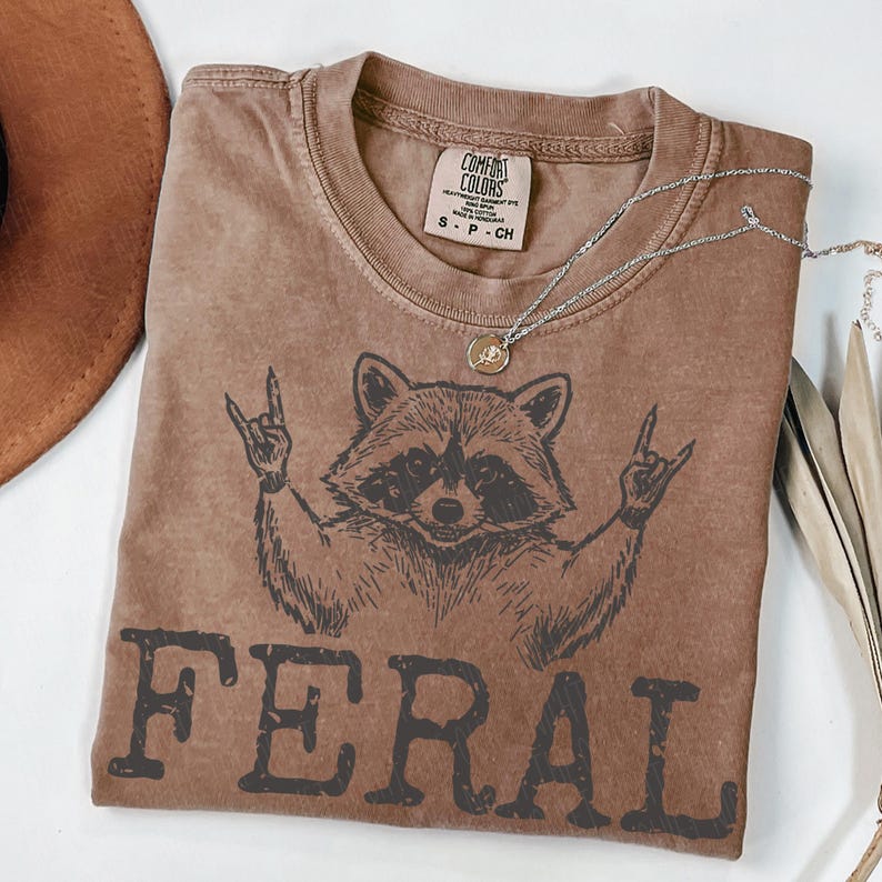 May include: Brown t-shirt with a raccoon graphic making a rock and roll hand gesture. The word "FERAL" is printed in bold letters below the raccoon. A silver necklace with a pendant is draped across the shirt.