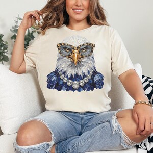 May include: A cream-colored short-sleeve t-shirt featuring a graphic of a bald eagle wearing leopard-print glasses and a silver-toned crystal necklace. The eagle has white head feathers and dark blue chest feathers, rendered in a textured paint style.