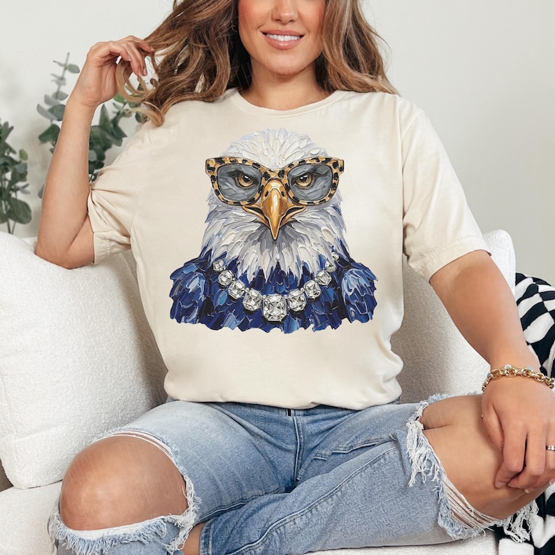 May include: A cream-colored short-sleeve t-shirt featuring a graphic of a bald eagle wearing leopard-print glasses and a silver-toned crystal necklace. The eagle has white head feathers and dark blue chest feathers, rendered in a textured paint style.