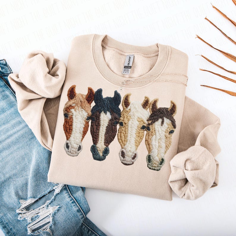 Peut inclure: Sweat-shirt beige avec un motif brod&eacute; de quatre t&ecirc;tes de chevaux dans les tons marron, noir et cr&egrave;me. Le sweat-shirt est pli&eacute;, avec les manches et une paire de jeans bleus visibles en arri&egrave;re-plan. Le motif est centr&eacute; sur la poitrine.