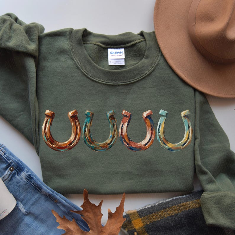 May include: Olive green sweatshirt featuring a row of five colorful horseshoe designs. The horseshoes are painted with shades of brown, orange, blue, and yellow. The sweatshirt has a crew neck and long sleeves, perfect for casual wear.