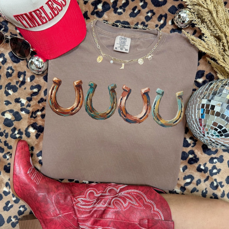 May include: A brown t-shirt with five horseshoe graphics in shades of brown, blue, and orange. The shirt is paired with a red baseball cap with the word "TIMELESS" and red cowboy boots. A gold necklace and sunglasses are also visible.