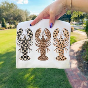 May include: A white t-shirt featuring three lobster designs. The lobsters have different patterns: black and white checkered, leopard print, and tan with black spots. The shirt is held up against a green grassy background.