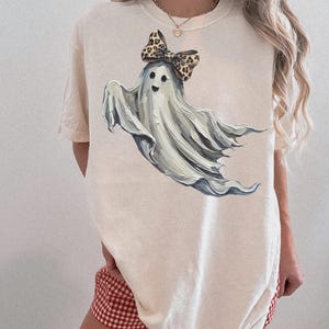 May include: A cream-coloured t-shirt with a playful ghost illustration. The ghost has a cheerful face and a leopard print bow. The design uses shades of white and grey, giving it a soft, artistic appearance. A fun, Halloween-themed garment.
