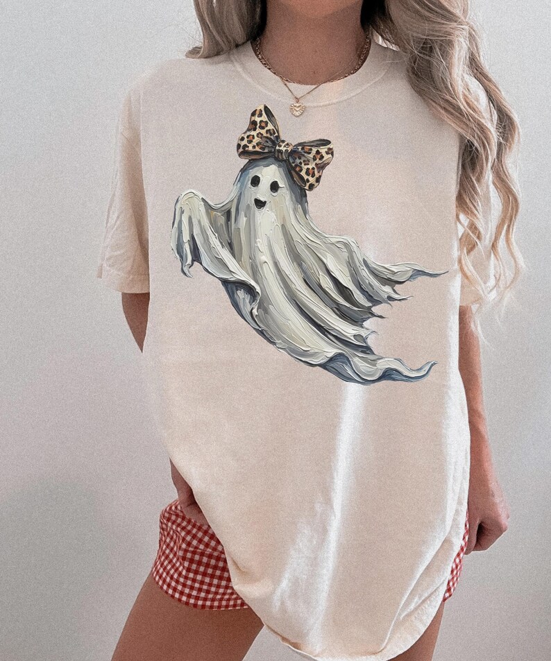 May include: A cream-coloured t-shirt with a playful ghost illustration. The ghost has a cheerful face and a leopard print bow. The design uses shades of white and grey, giving it a soft, artistic appearance. A fun, Halloween-themed garment.