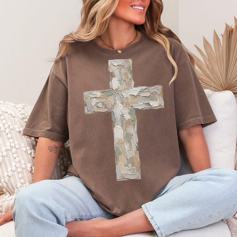 May include: A brown t-shirt featuring a large, textured cross design in shades of white, green, and beige. The shirt has short sleeves and a relaxed fit. The person is wearing light blue jeans.