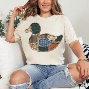 May include: A cream-colored short-sleeve t-shirt featuring a large duck graphic on the front. The duck is composed of various fabric patches in shades of green, brown, blue, and tan, showcasing different textures like corduroy and plaid patterns.
