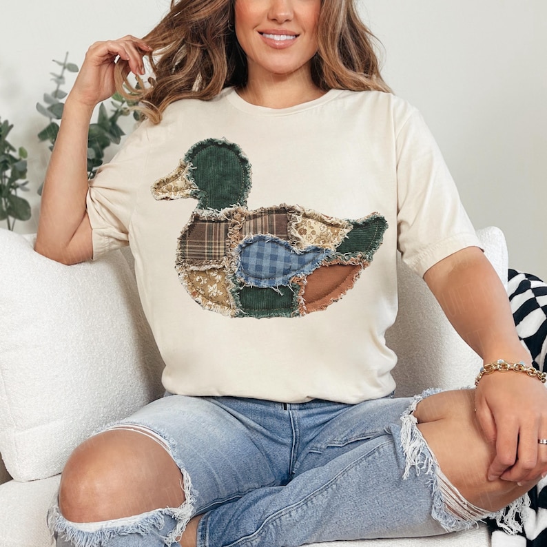 May include: A cream-colored short-sleeve t-shirt featuring a large duck graphic on the front. The duck is composed of various fabric patches in shades of green, brown, blue, and tan, showcasing different textures like corduroy and plaid patterns.
