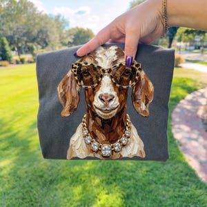 May include: A dark gray t-shirt featuring a painterly illustration of a brown and white goat wearing leopard print glasses and a gold chain necklace with clear square crystals. The goat has textured brushstrokes and a neutral facial expression.
