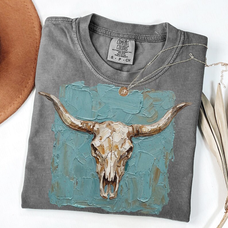 May include: A grey Comfort Colors t-shirt with a painted bull skull design on a blue background. The skull is detailed with tan and white tones, and the shirt is accessorized with a silver necklace. A brown hat is visible in the background.