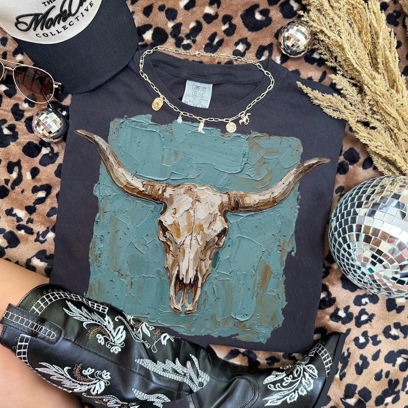 May include: A dark grey t-shirt with a textured bull skull design on a teal background. The image includes a black cap with white text, sunglasses, a gold necklace, a disco ball, and black cowboy boots with white embroidery.