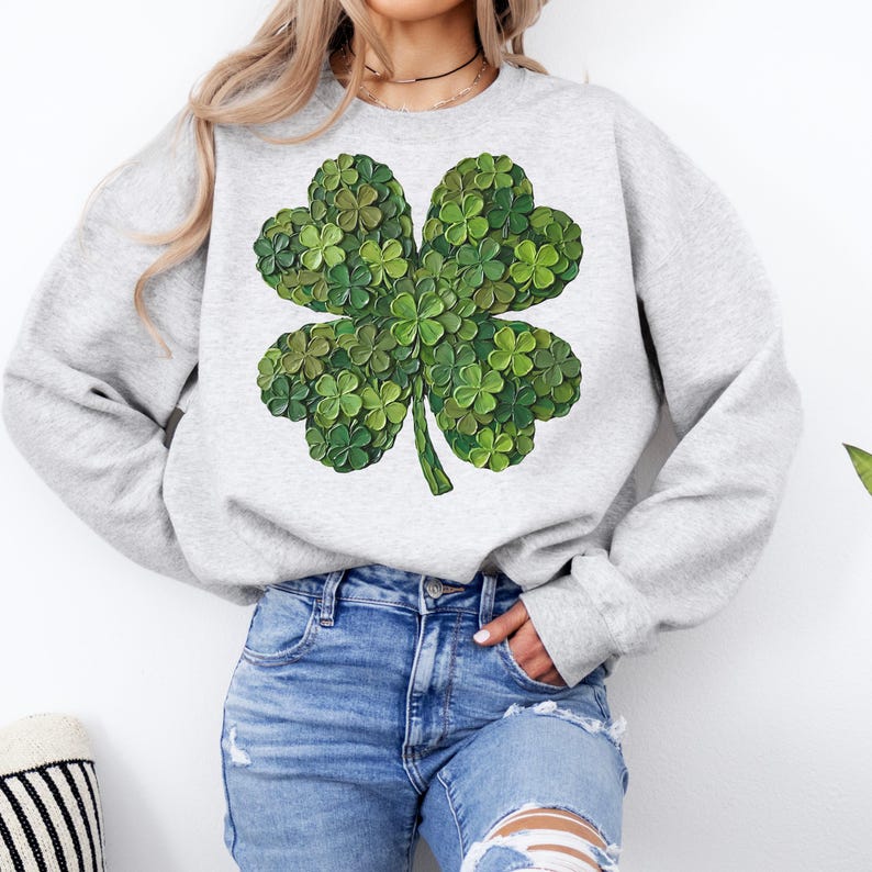 May include: A light gray sweatshirt featuring a large, detailed four-leaf clover design. The clover is composed of many small, textured green leaves, creating a dimensional effect. The sweatshirt has long sleeves and a crew neck.