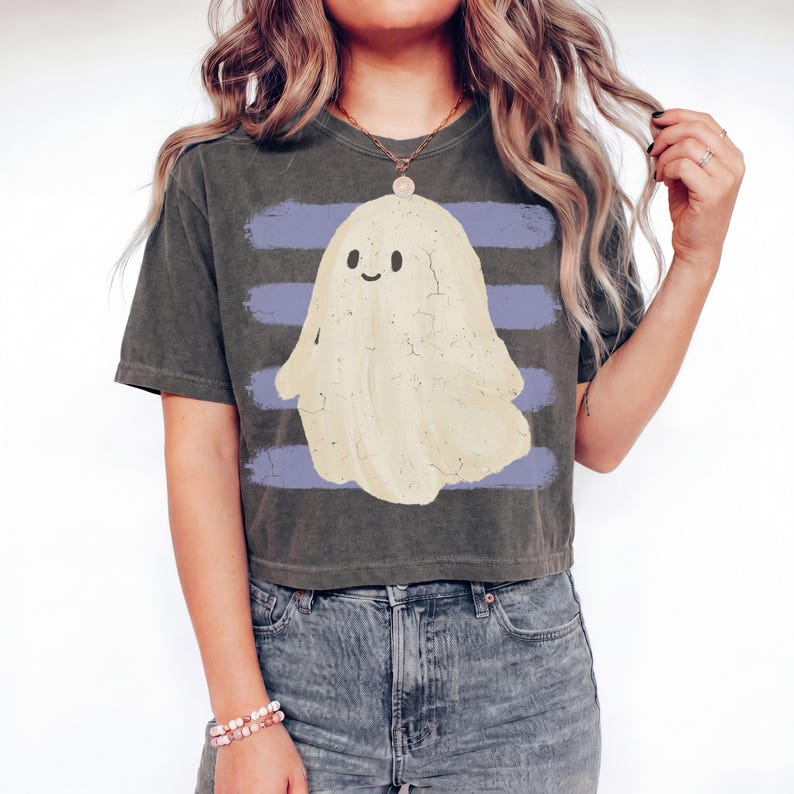 May include: A charcoal gray short-sleeve t-shirt featuring a central graphic of a cream-colored ghost with a smiling face. The ghost is positioned over three horizontal light purple stripes. The shirt has a vintage-style washed-out fabric texture.
