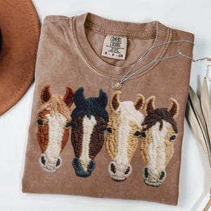 Peut inclure: Un t-shirt Comfort Colors marron avec des t&ecirc;tes de chevaux brod&eacute;es en marron, beige et noir. Un collier argent&eacute; avec un pendentif est pos&eacute; sur le t-shirt. Le t-shirt est de taille small.