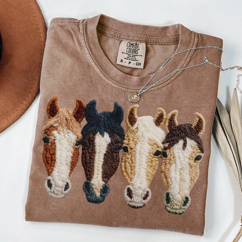 Peut inclure: Un t-shirt Comfort Colors marron avec des t&ecirc;tes de chevaux brod&eacute;es en marron, beige et noir. Un collier argent&eacute; avec un pendentif est pos&eacute; sur le t-shirt. Le t-shirt est de taille small.