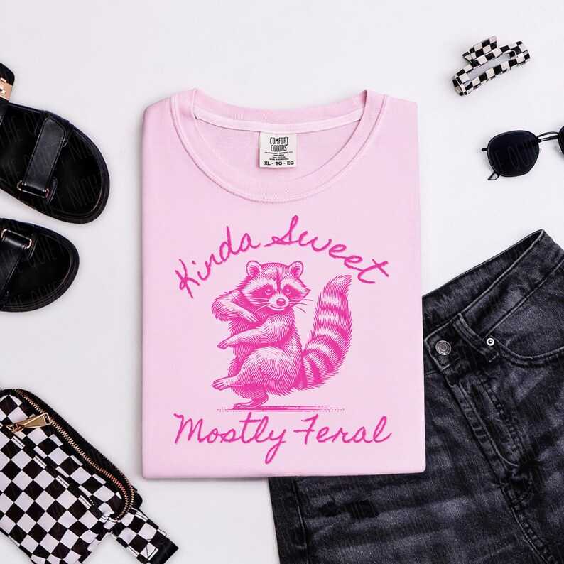 May include: A light pink sweatshirt with a raccoon graphic and the text "Kinda Sweet, Mostly Feral" in pink. The sweatshirt is surrounded by black sandals, sunglasses, a checkered bag, and black denim jeans.