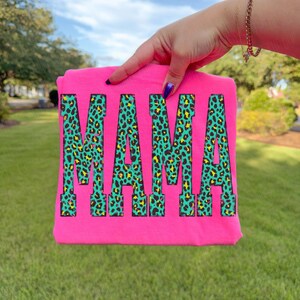 K&ouml;nnte beinhalten: Ein leuchtend neonpinkes T-Shirt mit dem Wort MAMA in einer gro&szlig;en Blockschrift. Die Buchstaben sind mit einem lebendigen Leopardenmuster in T&uuml;rkis, Gelb und Schwarz gef&uuml;llt. Das Kleidungsst&uuml;ck wird vor einem unscharfen gr&uuml;nen Rasen gehalten.