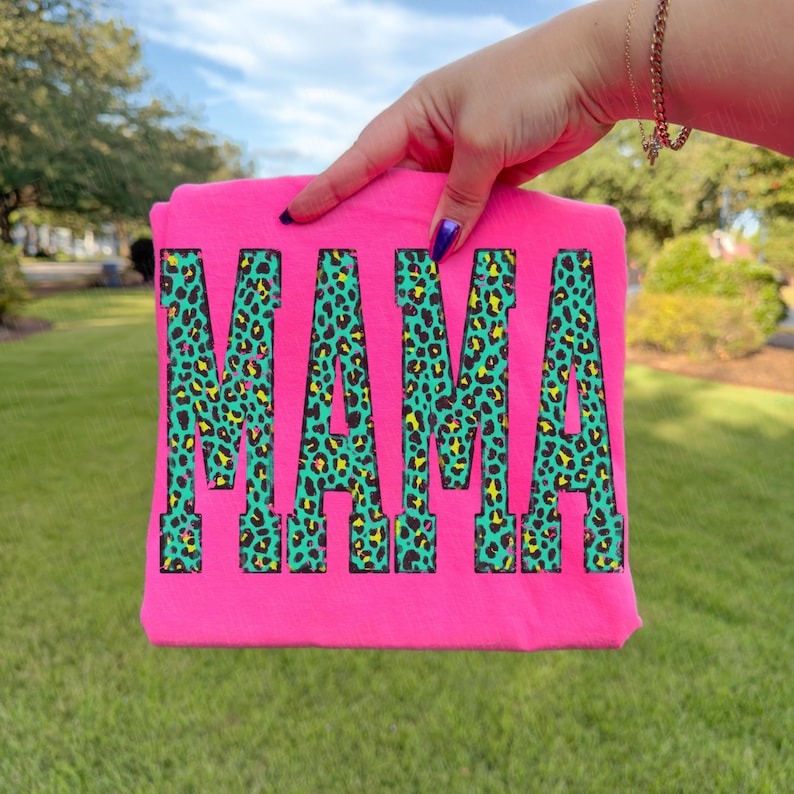 K&ouml;nnte beinhalten: Ein leuchtend neonpinkes T-Shirt mit dem Wort MAMA in einer gro&szlig;en Blockschrift. Die Buchstaben sind mit einem lebendigen Leopardenmuster in T&uuml;rkis, Gelb und Schwarz gef&uuml;llt. Das Kleidungsst&uuml;ck wird vor einem unscharfen gr&uuml;nen Rasen gehalten.