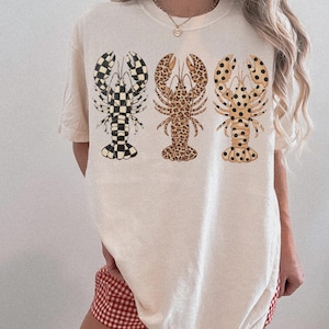 May include: Cream-colored t-shirt featuring three lobster designs. The lobsters have unique patterns: checkered, leopard print, and spotted. The shirt is a casual, oversized fit, suitable for everyday wear.