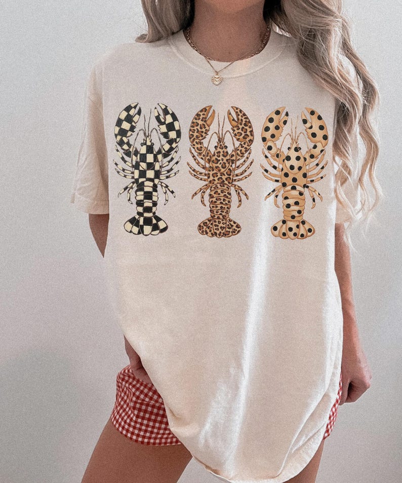 May include: Cream-colored t-shirt featuring three lobster designs. The lobsters have unique patterns: checkered, leopard print, and spotted. The shirt is a casual, oversized fit, suitable for everyday wear.