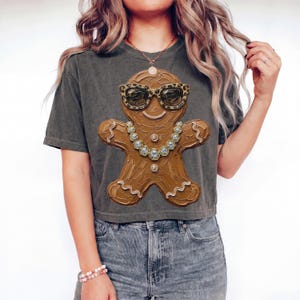 May include: A charcoal gray short-sleeve t-shirt featuring a large graphic of a brown gingerbread person wearing leopard print sunglasses and a silver-toned chain necklace with clear crystals. The gingerbread figure has a textured, painted appearance.