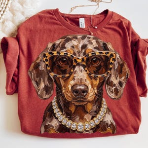 May include: A rust-colored t-shirt featuring a graphic of a dapple dachshund wearing leopard-print sunglasses and a chunky rhinestone collar. The dog illustration has a textured, painterly style with brown, tan, and black tones.