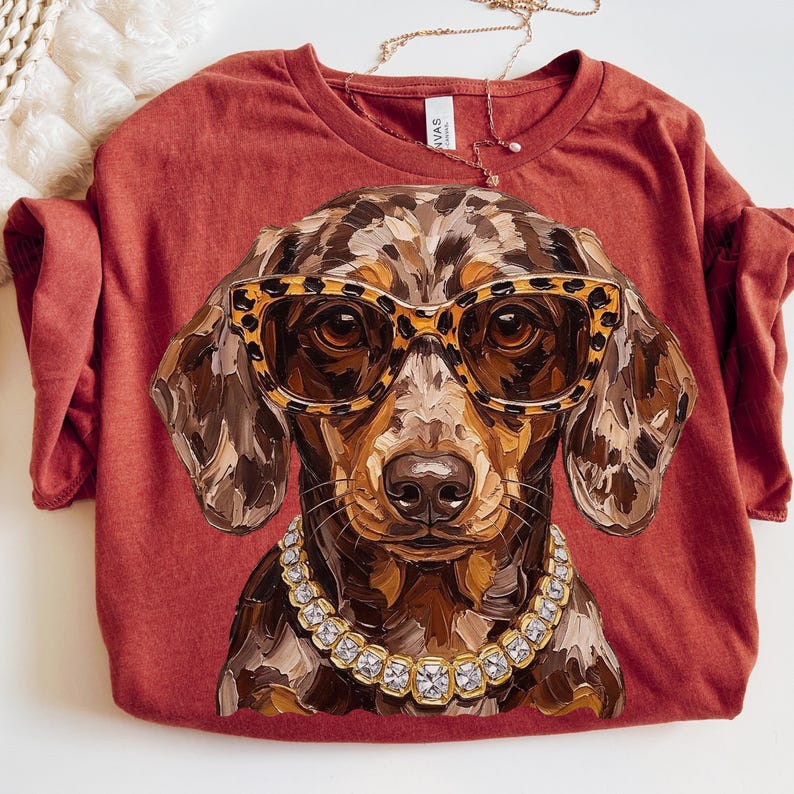 May include: A rust-colored t-shirt featuring a graphic of a dapple dachshund wearing leopard-print sunglasses and a chunky rhinestone collar. The dog illustration has a textured, painterly style with brown, tan, and black tones.