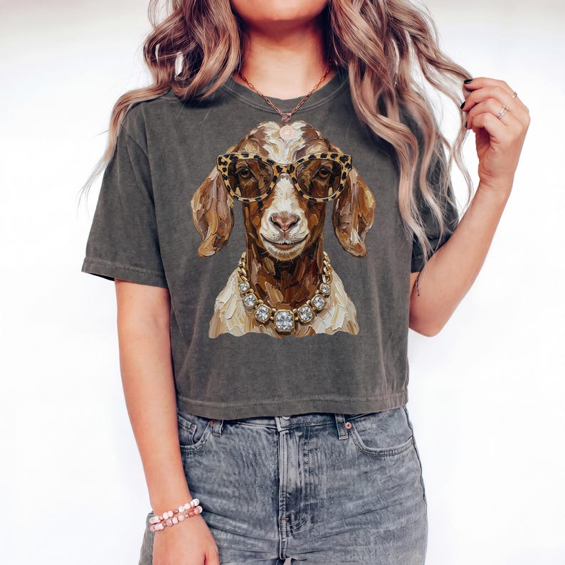 May include: A charcoal gray short-sleeve t-shirt featuring a graphic illustration of a brown goat wearing leopard print glasses and a chunky gold-toned necklace with large clear crystals. The goat is depicted in a painterly style with visible brushstrokes.