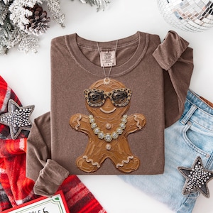 May include: A brown long-sleeve shirt featuring a graphic of a gingerbread person wearing leopard print sunglasses and a crystal-style necklace. The shirt has a crew neckline and a Comfort Colors brand label printed on the inside of the collar.
