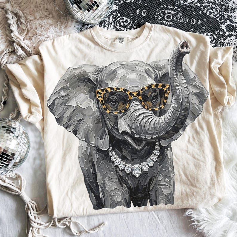 May include: A cream-colored t-shirt featuring a graphic of a gray elephant with a textured, impasto-style paint effect. The elephant wears leopard-print glasses and a silver-toned necklace with clear, faceted stones around its neck.