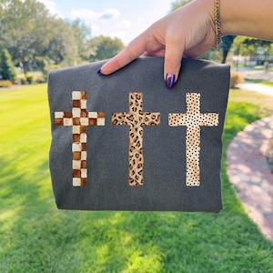 May include: Dark gray fabric featuring three crosses with unique patterns. One cross has a brown and white checkered design, another has a leopard print, and the third has a dotted pattern. The crosses are centered on the fabric.