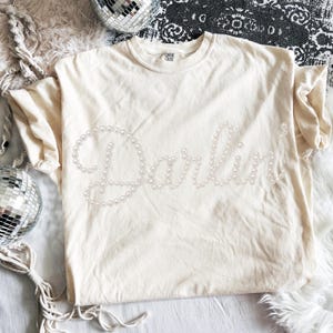 Pu&ograve; includere: T-shirt color crema con la scritta "Darling" realizzata con perle finte. La maglietta ha maniche corte e girocollo. La maglietta &egrave; appoggiata su una superficie bianca con palle da discoteca sullo sfondo.