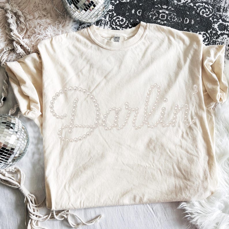 Pu&ograve; includere: T-shirt color crema con la scritta "Darling" realizzata con perle finte. La maglietta ha maniche corte e girocollo. La maglietta &egrave; appoggiata su una superficie bianca con palle da discoteca sullo sfondo.