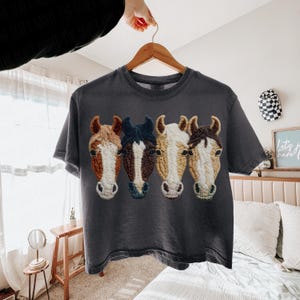 Peut inclure: T-shirt gris fonc&eacute; orn&eacute; de quatre t&ecirc;tes de chevaux brod&eacute;es en marron, blanc et bleu. Les t&ecirc;tes de chevaux sont dispos&eacute;es en rang&eacute;e sur le devant. Le t-shirt est suspendu &agrave; un cintre en bois.