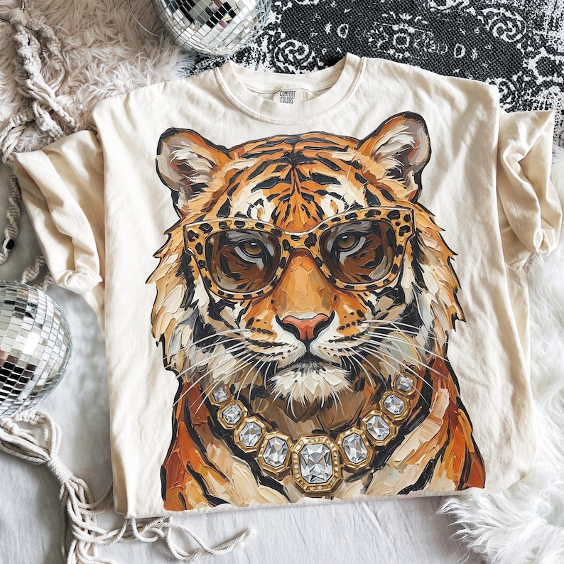 K&ouml;nnte beinhalten: Cremefarbenes T-Shirt mit einer detaillierten Illustration eines Tigers, der eine Sonnenbrille mit Leopardenmuster und eine Diamantkette tr&auml;gt. Das Fell des Tigers ist in Orange-, Braun- und Wei&szlig;t&ouml;nen gehalten. Das Shirt hat einen Rundhalsausschnitt und kurze &Auml;rmel.