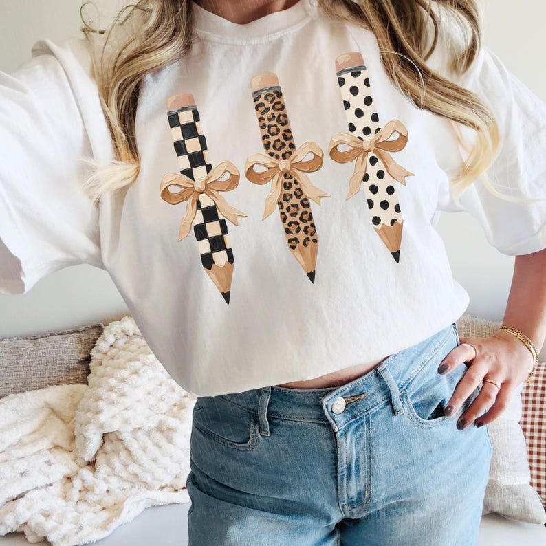 以下が含まれることがあります： 白のTシャツに、チェック柄、ヒョウ柄、水玉模様の3種類の装飾的な鉛筆が描かれています。各鉛筆にはベージュのリボンが付いています。鉛筆はTシャツに縦に配置されています。
