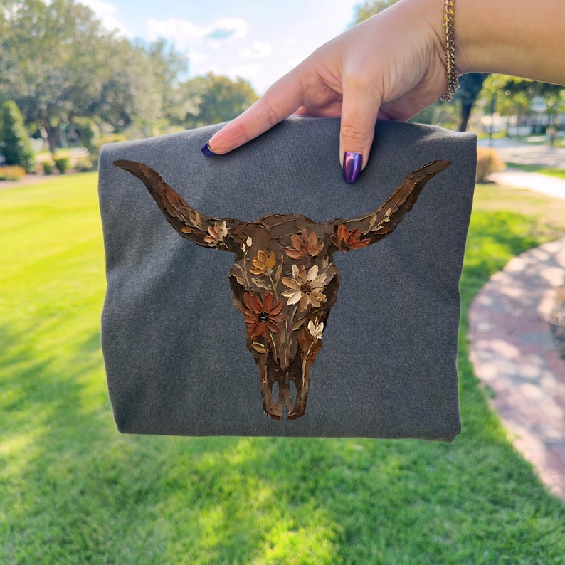 May include: A dark gray t-shirt featuring a detailed painting of a bull skull adorned with flowers. The artwork uses shades of brown, beige, and white, creating a rustic and artistic design. The shirt is held up against a green grassy background.