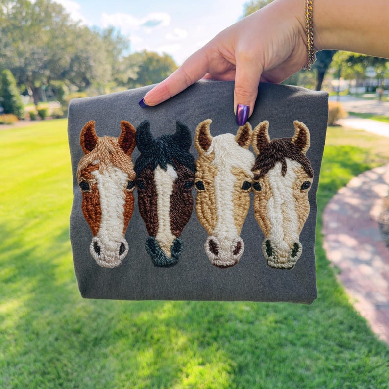 Peut inclure: Un article en tissu gris avec un motif brod&eacute; de quatre t&ecirc;tes de chevaux. Les chevaux sont de diff&eacute;rentes couleurs, notamment marron, noir et beige, avec des coutures d&eacute;taill&eacute;es. L'article est pr&eacute;sent&eacute; sur un fond vert.