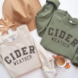 K&ouml;nnte beinhalten: Zwei Sweatshirts, eines in Creme und eines in Olivgr&uuml;n, mit dem Aufdruck "CIDER WEATHER" in einer verblichenen Schriftart im College-Stil. Ein beiger Fedora-Hut und ein Herbstblatt sind ebenfalls zu sehen. Eine kleine Kaffeetasse steht neben dem gr&uuml;nen Sweatshirt.