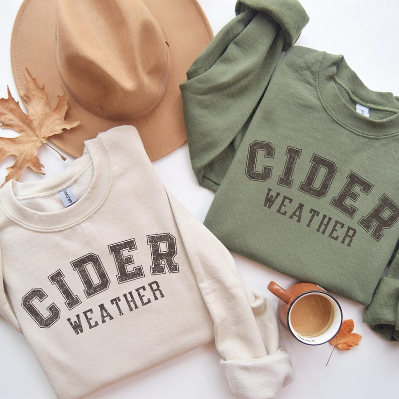 K&ouml;nnte beinhalten: Zwei Sweatshirts, eines in Creme und eines in Olivgr&uuml;n, mit dem Aufdruck "CIDER WEATHER" in einer verblichenen Schriftart im College-Stil. Ein beiger Fedora-Hut und ein Herbstblatt sind ebenfalls zu sehen. Eine kleine Kaffeetasse steht neben dem gr&uuml;nen Sweatshirt.