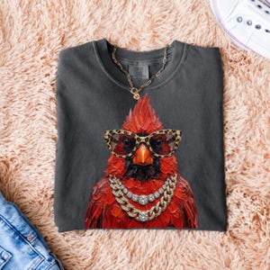 May include: Dark gray t-shirt featuring a red cardinal wearing leopard print glasses and a gold chain necklace with diamond accents. The bird is the focal point, with a detailed illustration. The shirt is folded on a pink, fluffy surface.