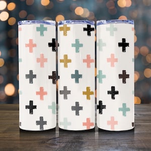 May include: Three white insulated tumblers with a repeating pattern of colorful crosses in shades of pink, green, yellow, gray, and black. The tumblers have a silver rim and a blue lid. The background is blurred with bokeh lights.