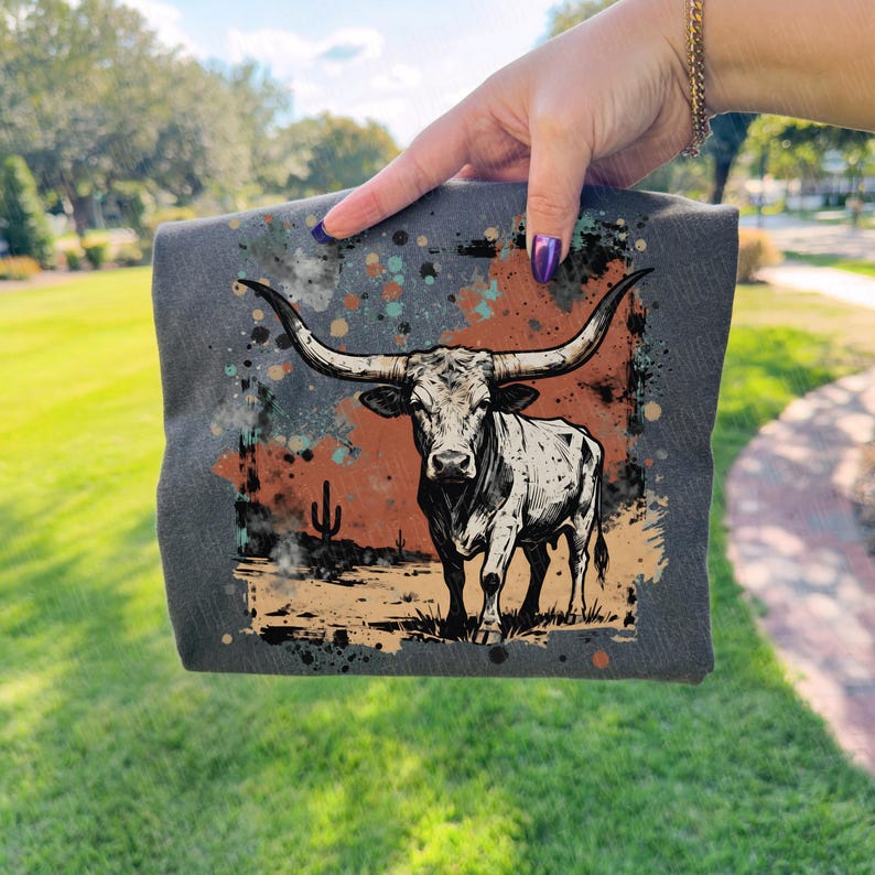 May include: A dark gray square textile featuring a black and white illustration of a longhorn steer. The design includes a desert landscape with a cactus and colorful paint splatters. The textile is held up by a hand.