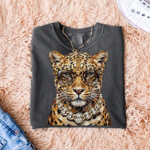 Puede incluir: Camiseta Comfort Colors gris oscuro con un leopardo que lleva gafas y un collar de diamantes. El leopardo tiene un patr&oacute;n de manchas doradas y marrones. Se ve una cadena dorada. La camiseta est&aacute; sobre una superficie rosa y esponjosa.