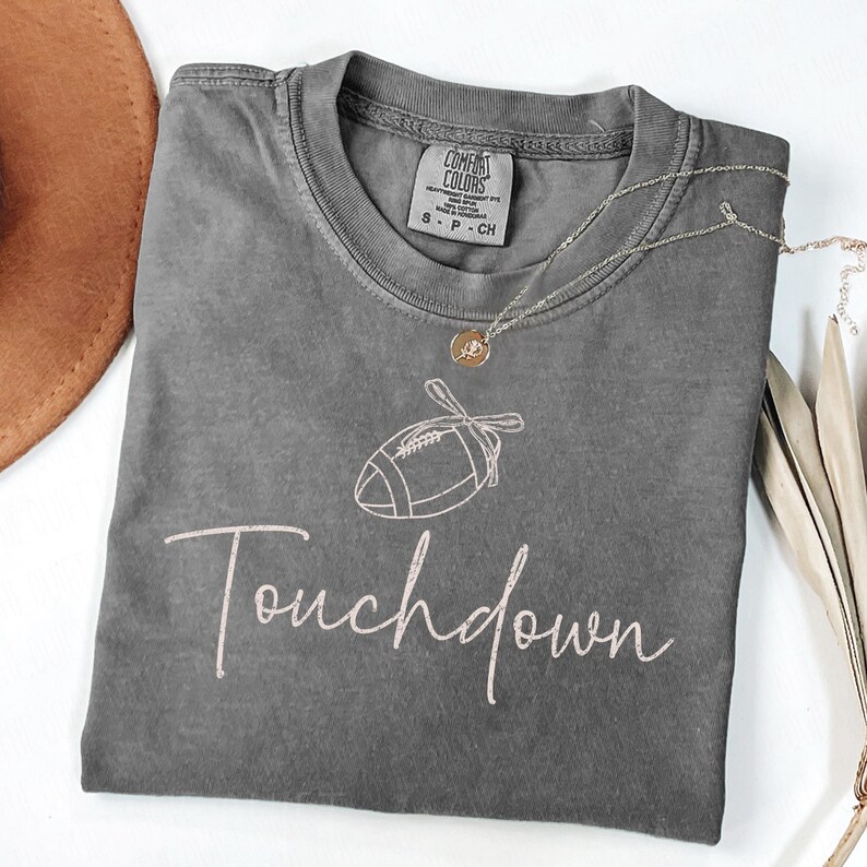 May include: A gray Comfort Colors t-shirt with a football graphic and the word "Touchdown" in a cursive font. A gold necklace with a pendant is draped across the shirt. The shirt is folded on a white surface.