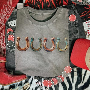 May include: A gray crew neck t-shirt with five horseshoe designs in shades of brown, blue, and orange. The shirt is surrounded by denim shorts, a red baseball cap, poker chips, dice, and a red bandana.
